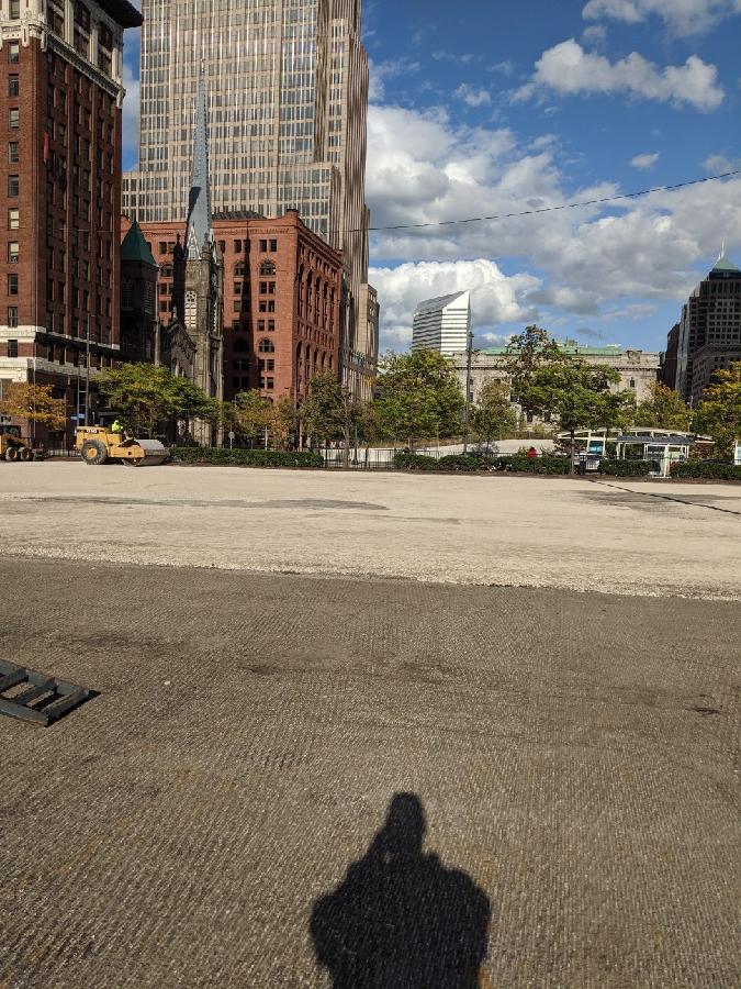 A large empty lot in a city with tall buildings, trees, and a blue sky. A person's shadow is cast on the lot.
