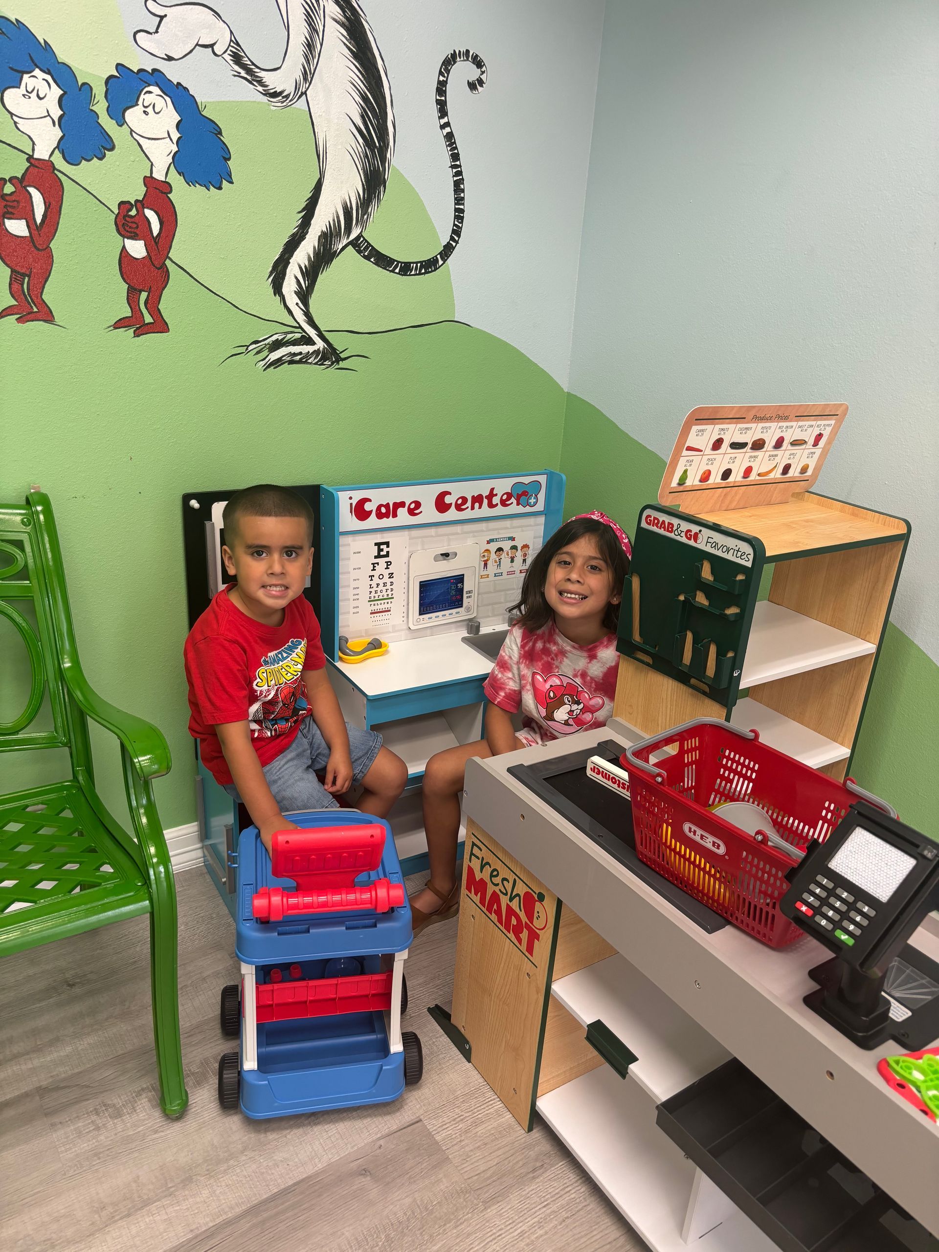 Two children playing at a Dr. Seuss-themed play grocery store. Boy holding a toy cart, girl smiling.