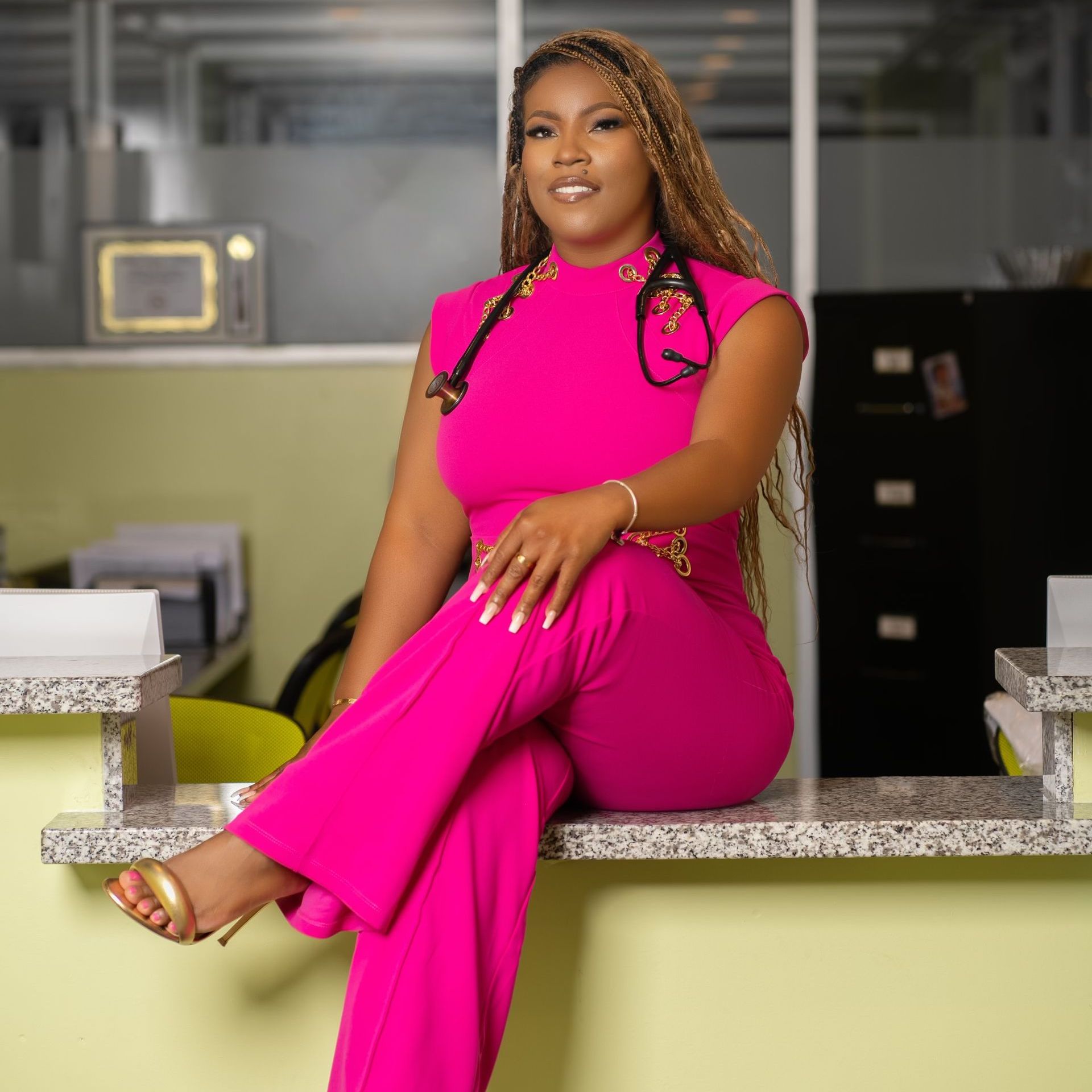 Woman in pink jumpsuit, stethoscope around neck, sitting on counter in office.
