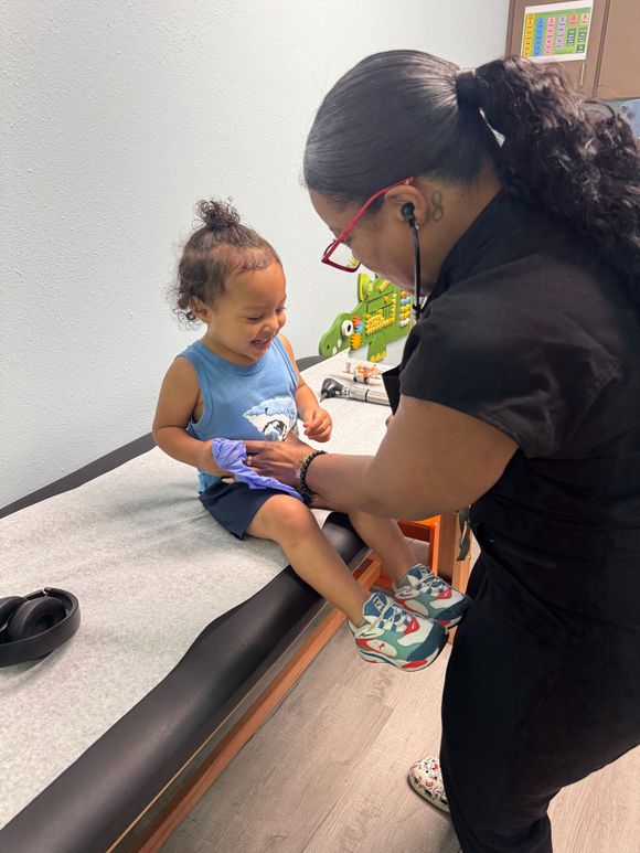 A smiling toddler's leg is examined by a woman with a stethoscope in a medical setting.