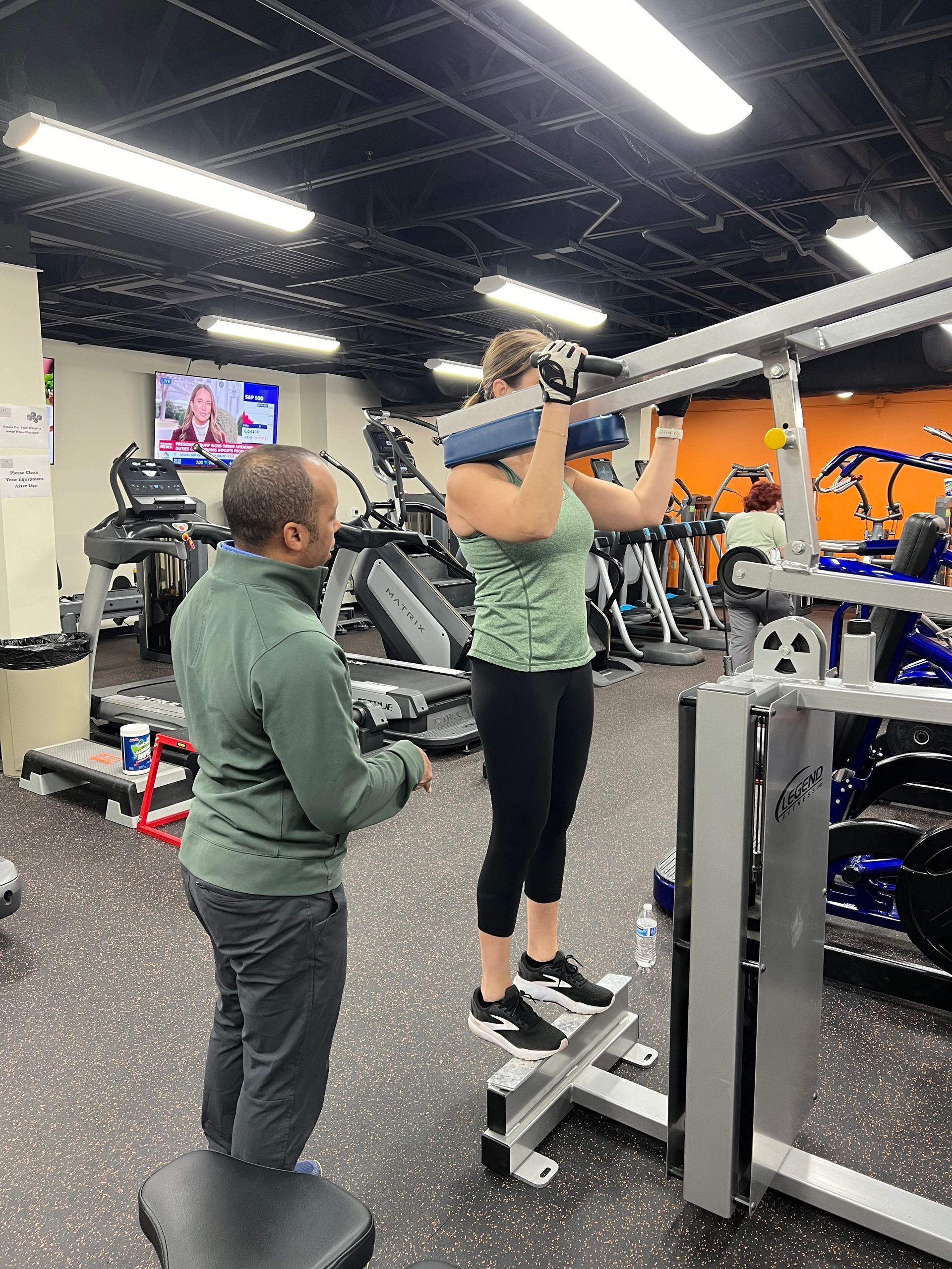 A man is standing next to a woman doing exercises in a gym.