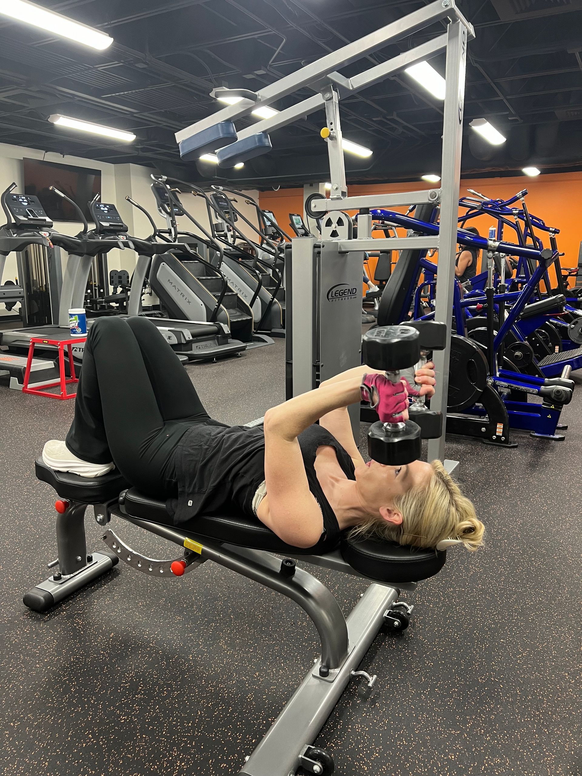 A woman is laying on a bench in a gym holding a dumbbell.