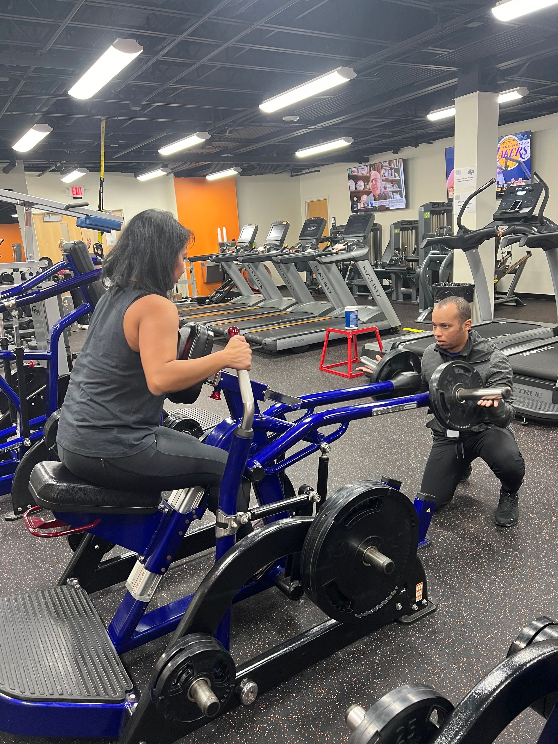 A woman is sitting on a machine in a gym while a man squats next to her.