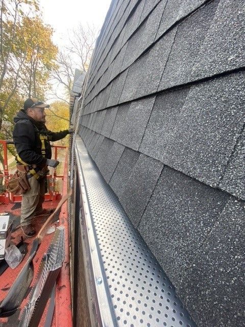 A worker installs gutter guard on a roof, wearing a safety harness.