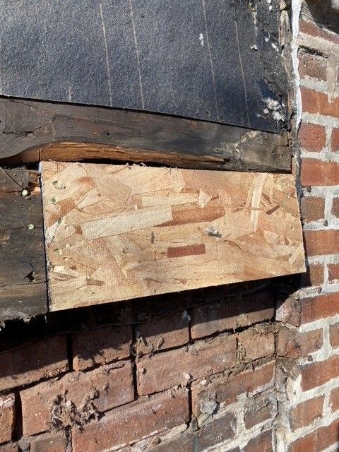 OSB patch over brick, likely a repair. Dark roofing material above, and visible brickwork below.