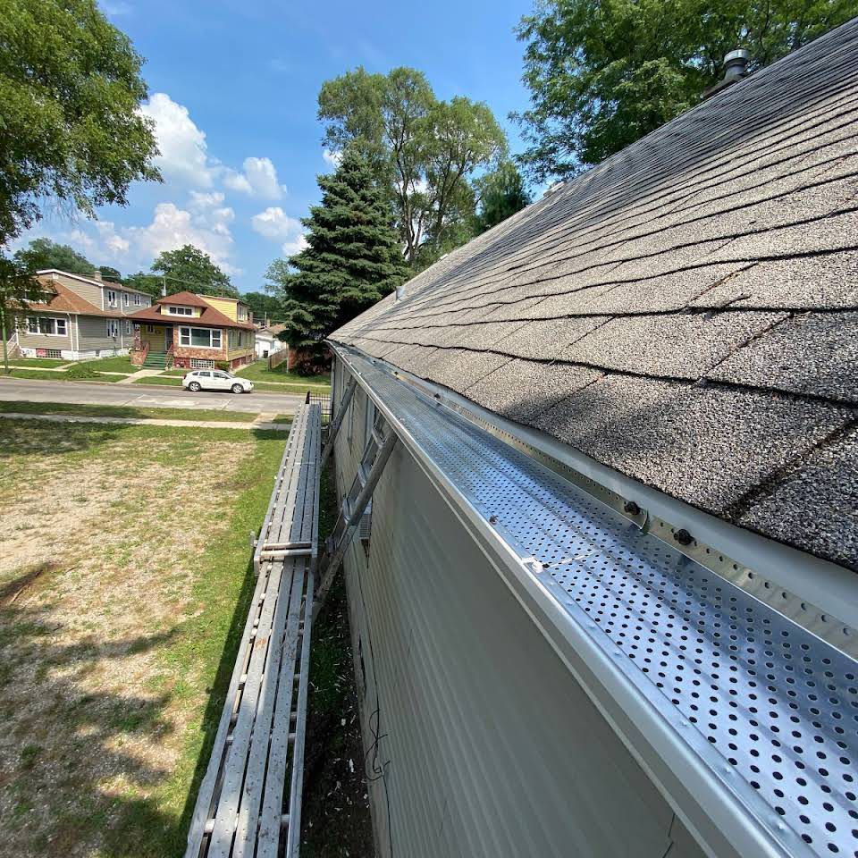 View of a house's roof and gutter with a leaf guard, street and houses in background on a sunny day.