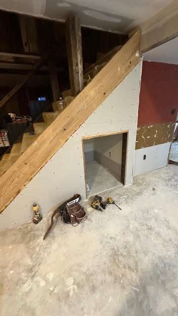 Staircase under construction with a small doorway. Tools sit on the concrete floor. Drywall and wood are visible.