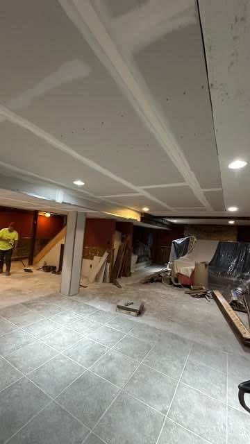 Basement renovation in progress. Drywall installed on ceiling, exposed beams, gray tiled floor, and construction materials scattered.