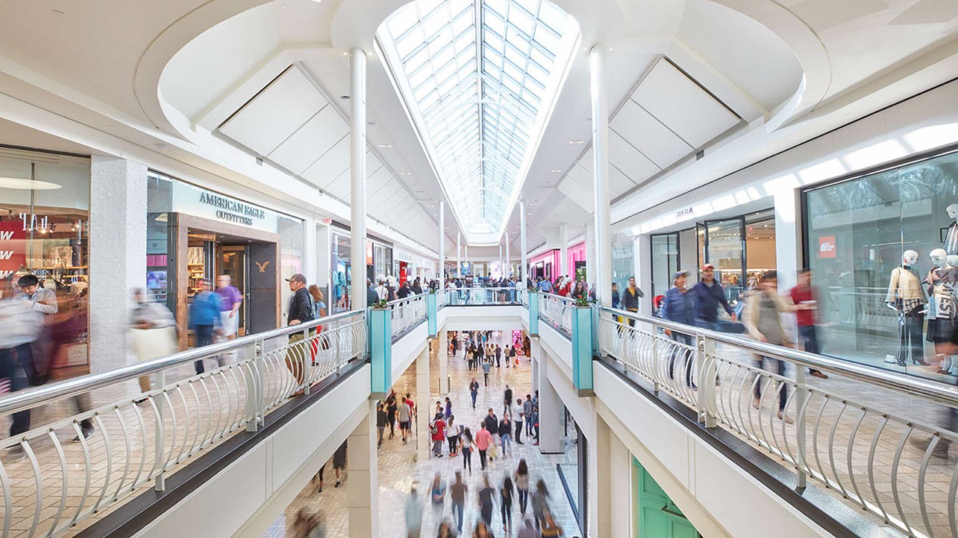 Interior view of a busy shopping mall with two levels of shops, glass ceiling, and shoppers.