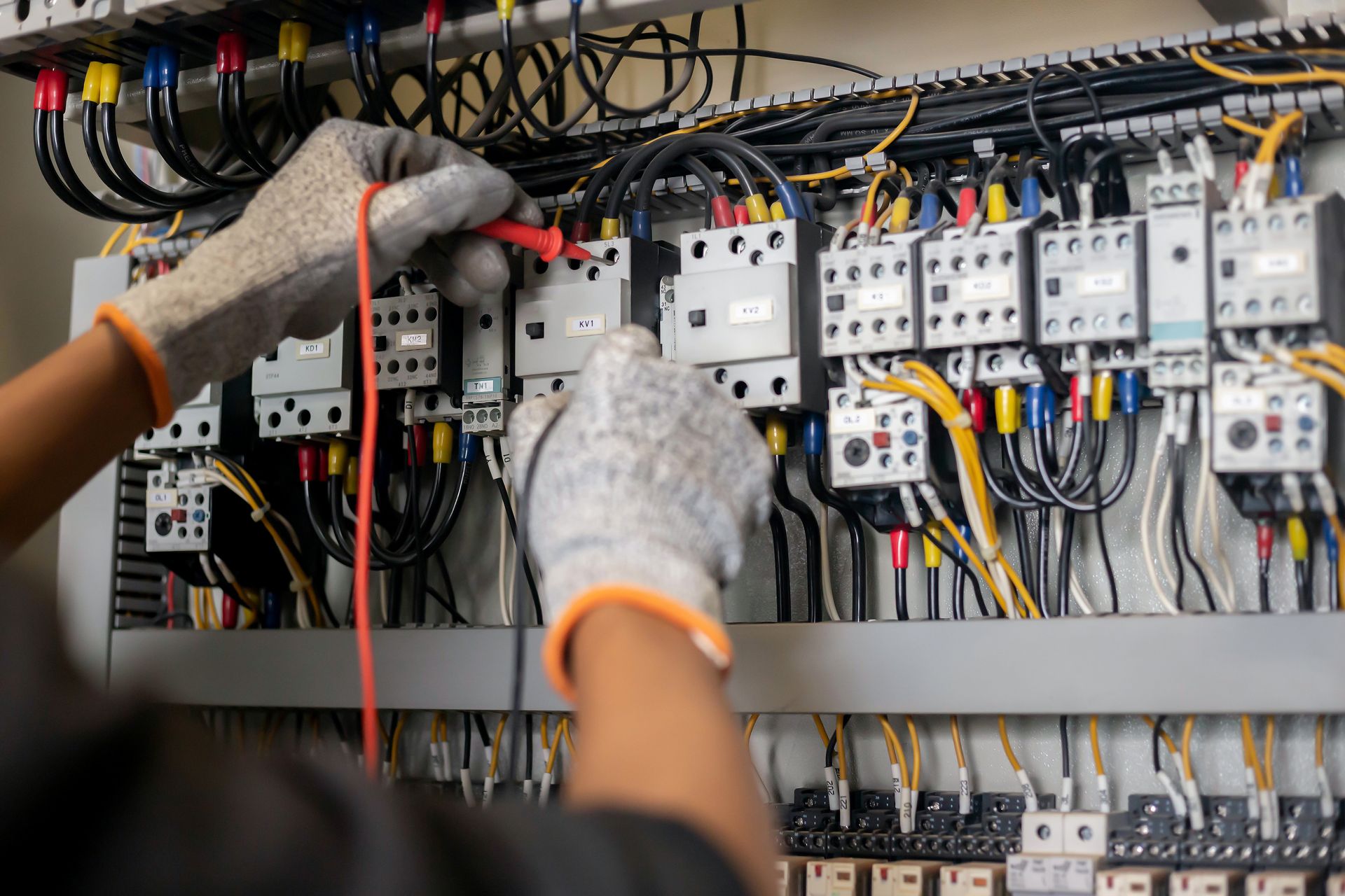 Person testing electrical panel with multimeter, wearing gloves. Wires are black, blue, yellow, and red.