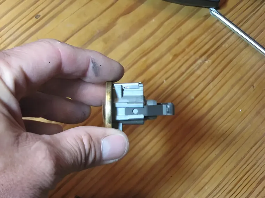 A hand holds a metallic door lock cylinder mechanism against a wooden table background.