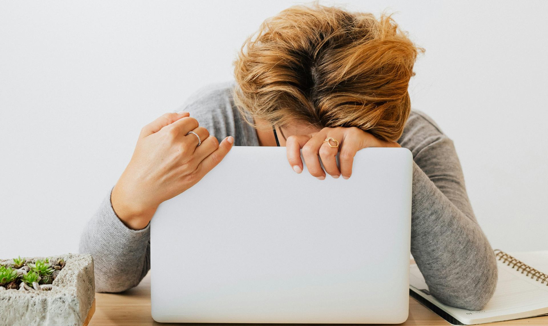 Woman with head down on a laptop, looking frustrated at a desk with a plant and notebook.