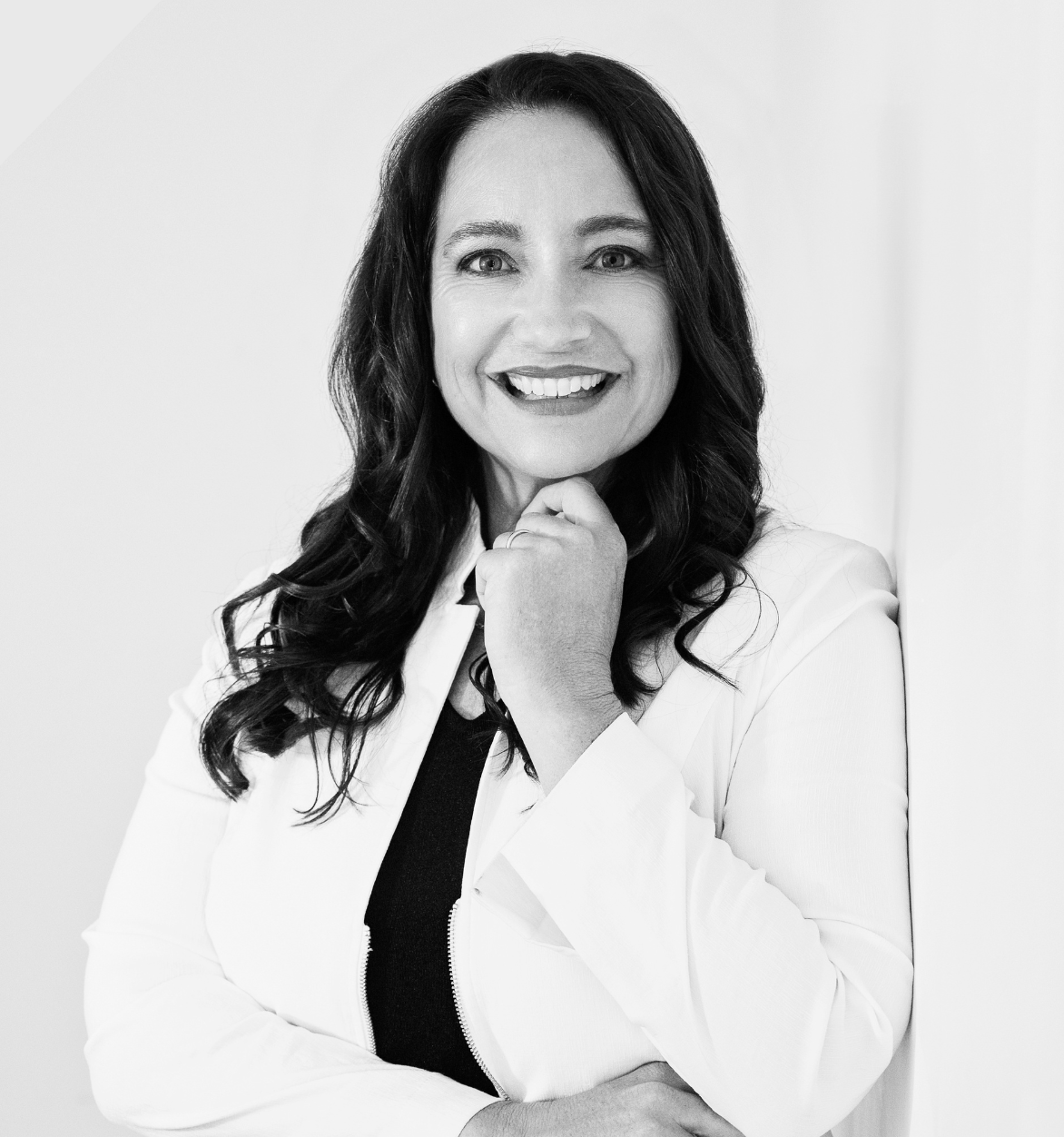 Woman in white blazer smiles, hand to chin, leaning against a white wall.