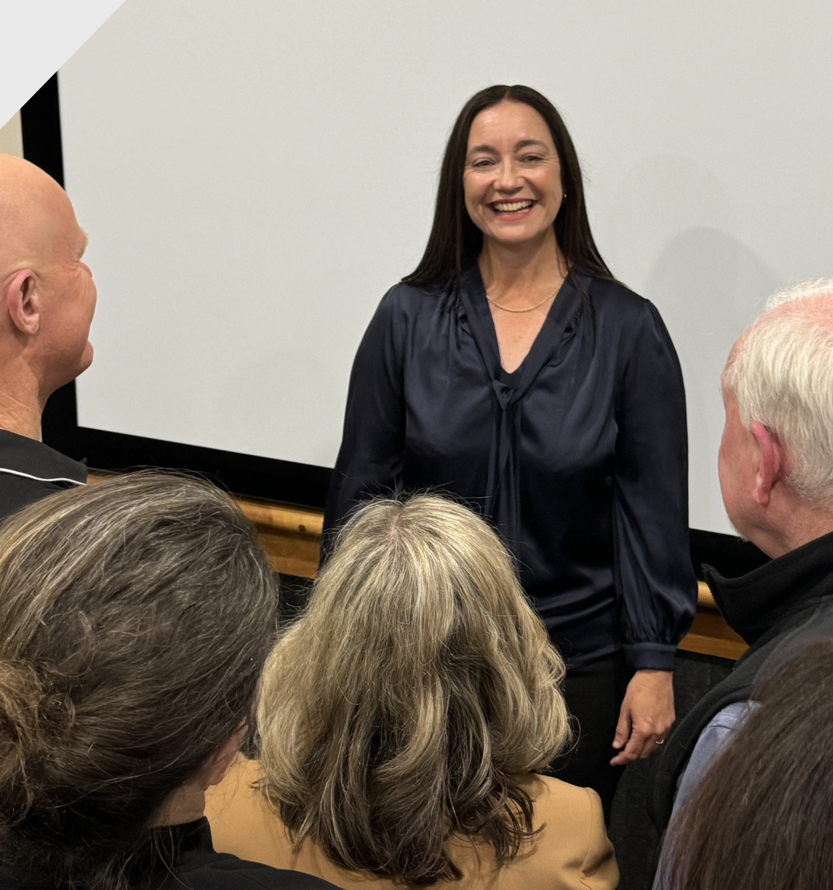 Woman in a dark blue blouse smiles, presenting to a group near a projector screen.