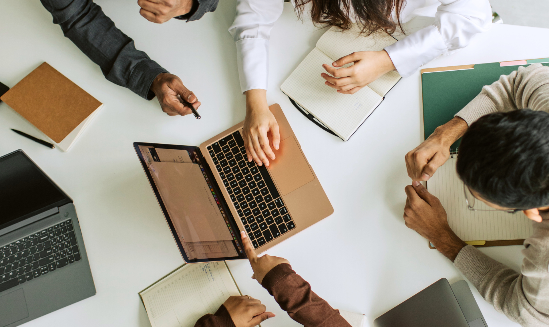 People gathered around a table with laptops, notebooks, and folders, collaborating and pointing at a screen.
