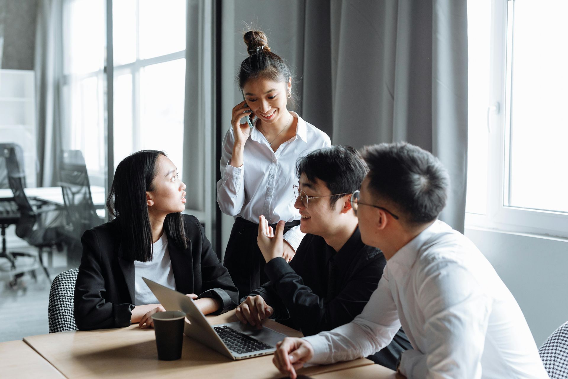 Four people in an office, one on the phone, collaborating around a laptop, discussing.