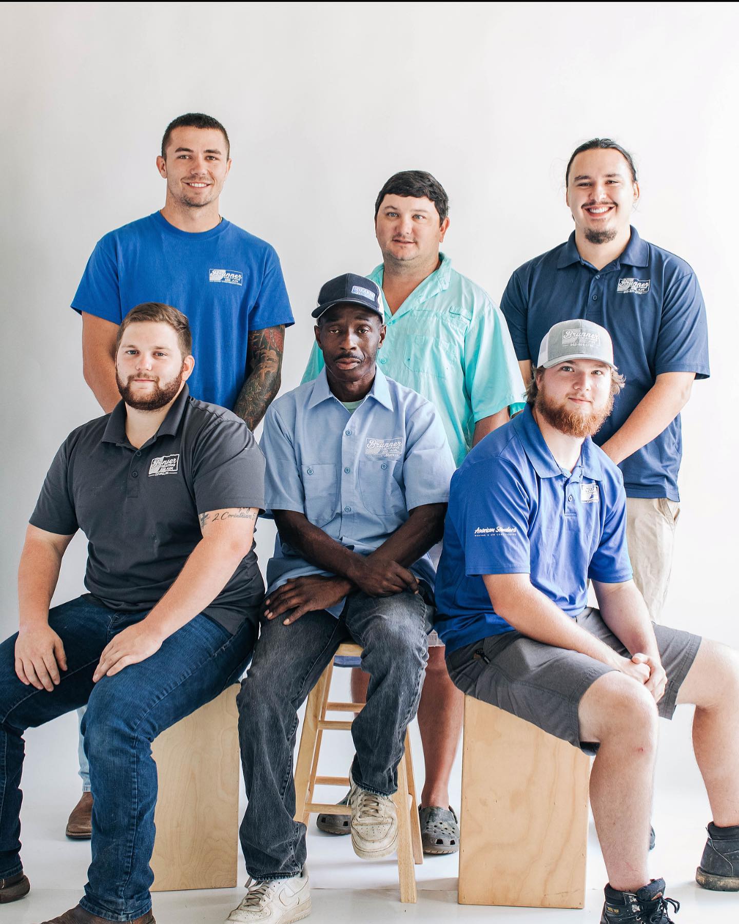Six individuals in branded work shirts pose for a group photo against a plain white background, three seated, three standing.