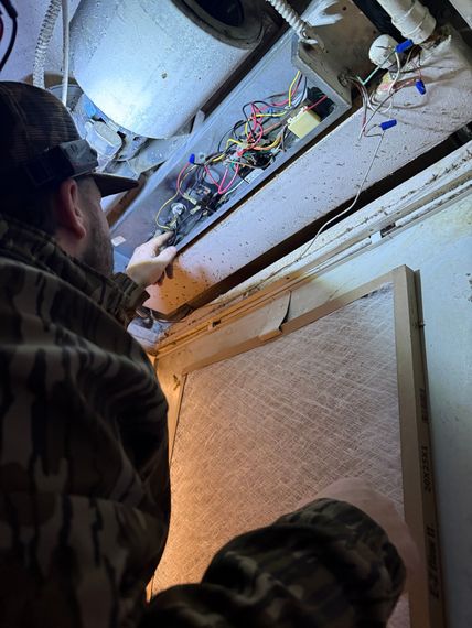 A person wearing camouflage clothing inspecting the electrical wiring inside an open HVAC unit ceiling panel.