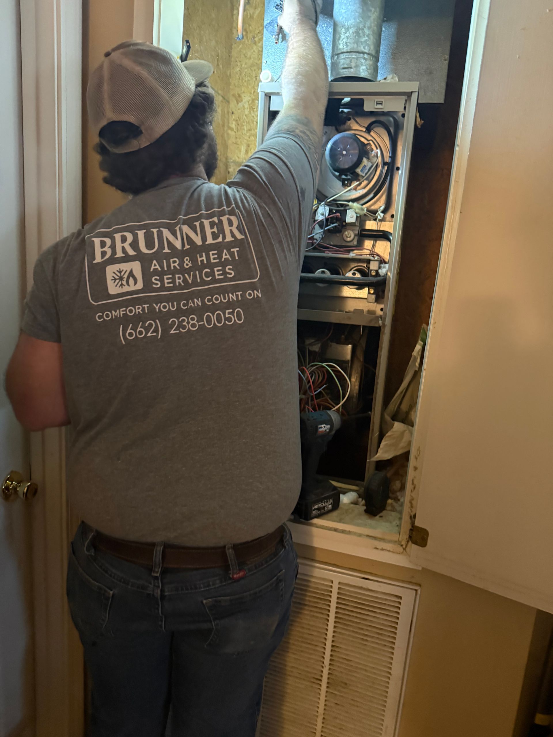 A service technician in a gray shirt works on the internal components of an open HVAC unit in a wall alcove.