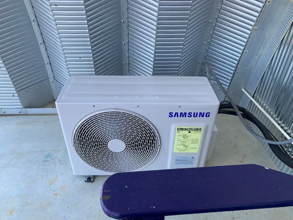 A white Samsung outdoor air conditioner unit positioned on a concrete floor against a corrugated metal wall.