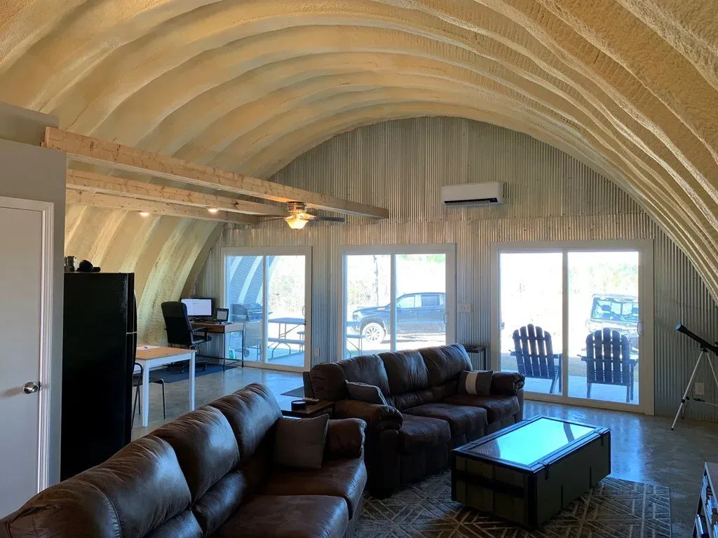 A spacious living room inside a Quonset hut style home with vaulted, insulated ceilings, leather furniture, and patio doors.