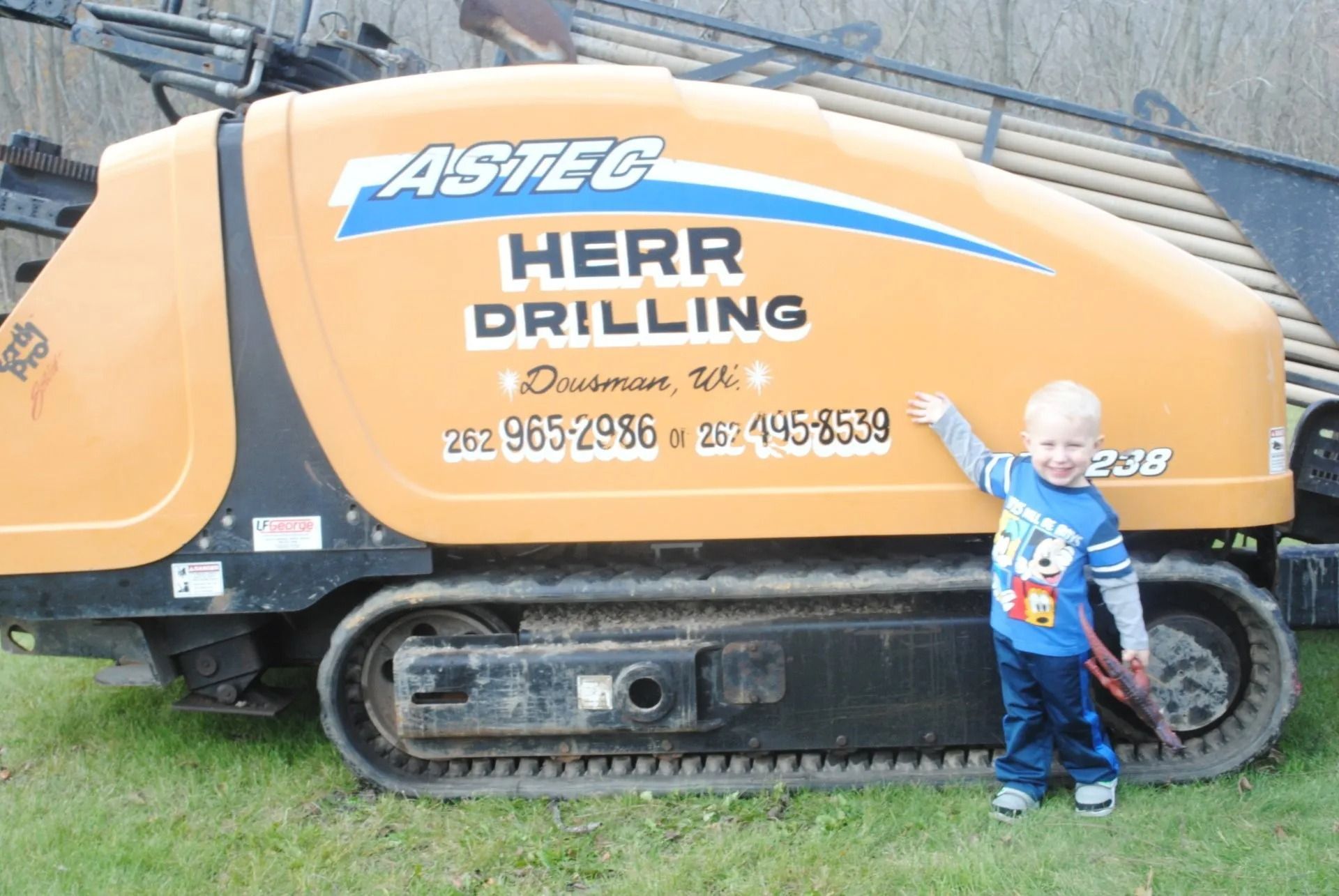 Child pointing at an Astec Herr Drilling machine in a grassy area. The machine is orange and black.