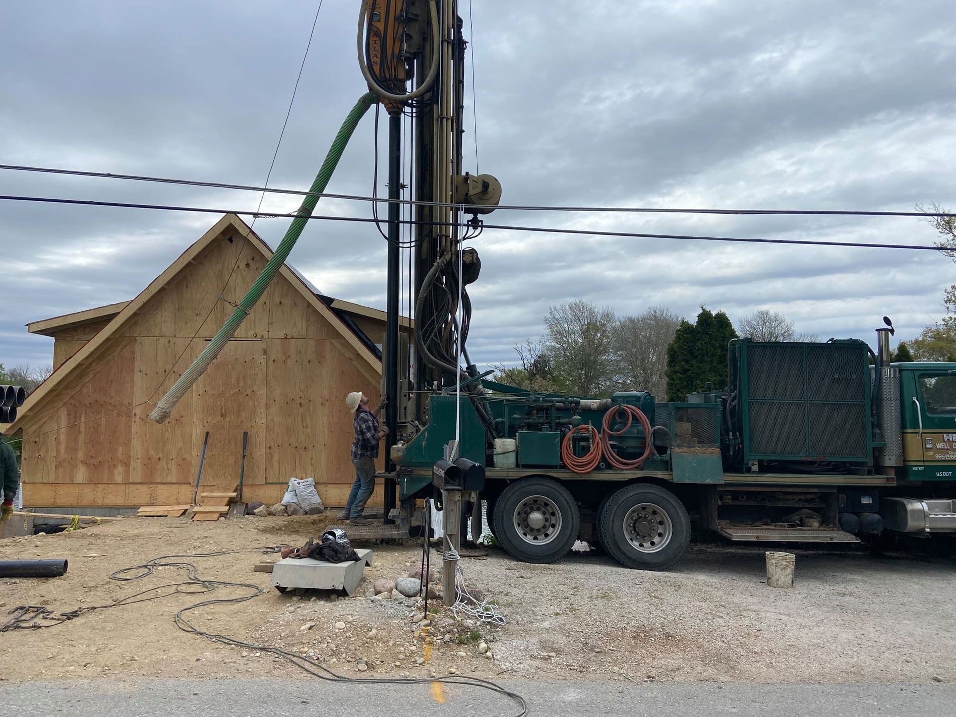 Drilling rig outside a wood-framed building; worker near rig, overcast sky.