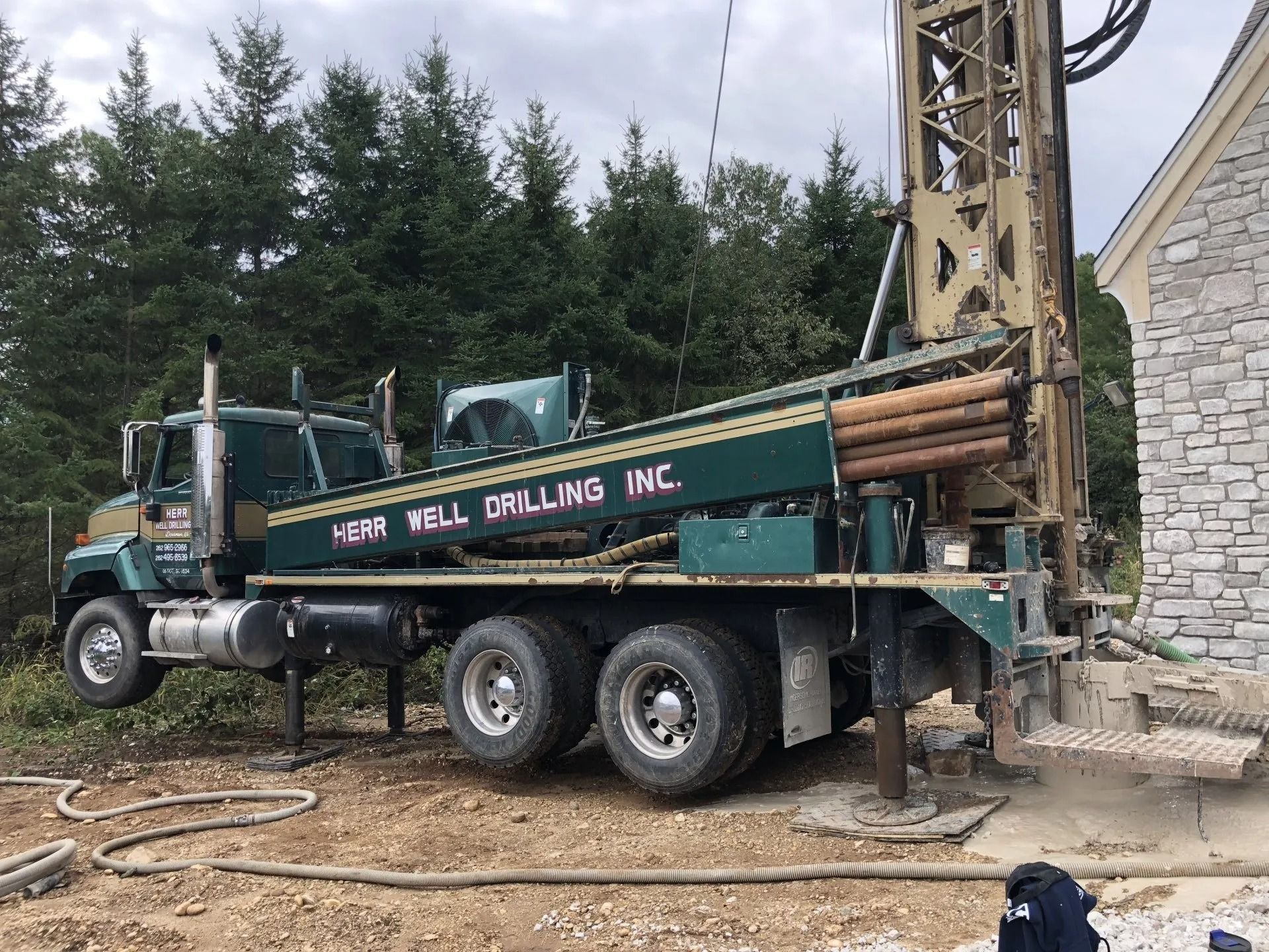 Green well drilling truck with a drilling rig operating near a house.