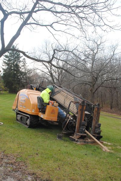 Person operating orange drilling machine on grassy ground. Bare trees in background.