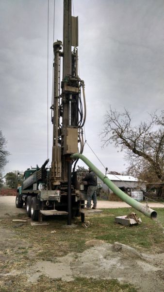 Well drilling rig on a truck in a grassy area, overcast sky. A person works near the rig.