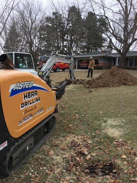 Drilling equipment in a yard, drilling crew, house in the background. Brown and white dirt pile.