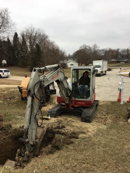 A person operates an excavator in a residential yard, digging a hole. Gray and red machine, cloudy sky.