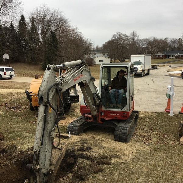Man operating an excavator in a residential area. Ground is dug up. A white truck and a tree stump grinder are visible.