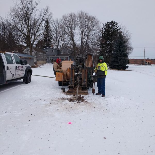 Two workers using drilling equipment in a snowy area, near a truck, houses, and trees.