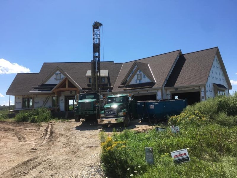 A well-drilling truck with an extended rig parked in front of a new, partially finished house under construction.