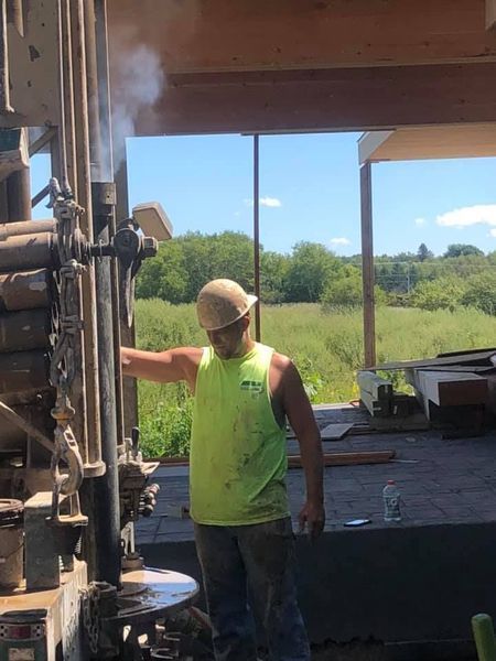 Man operating drilling equipment outdoors, wearing safety gear and neon green shirt. Smoke visible.