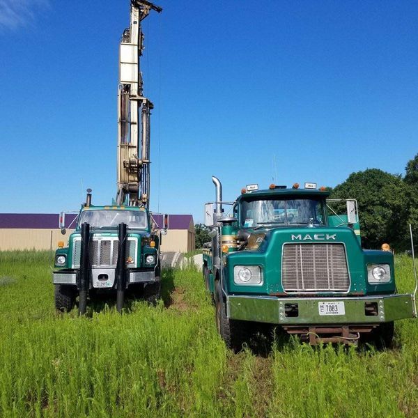 Two green Mack trucks in a grassy field; one has a tall drilling rig attached. Bright blue sky overhead.