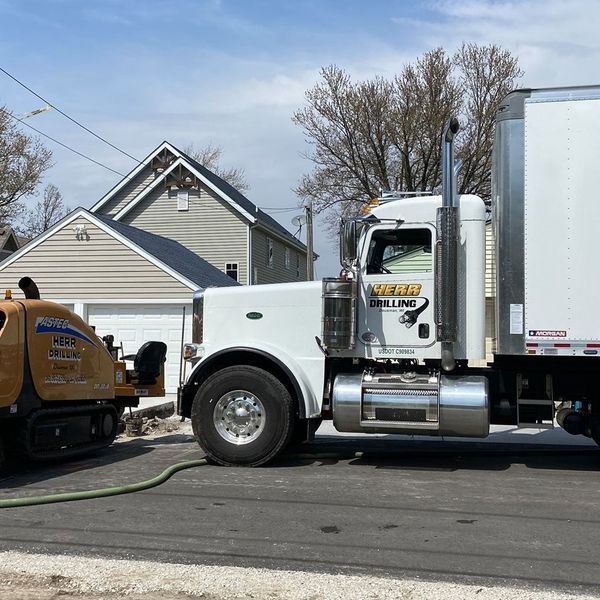 White semi-truck parked on a street next to a small construction machine, near a house.