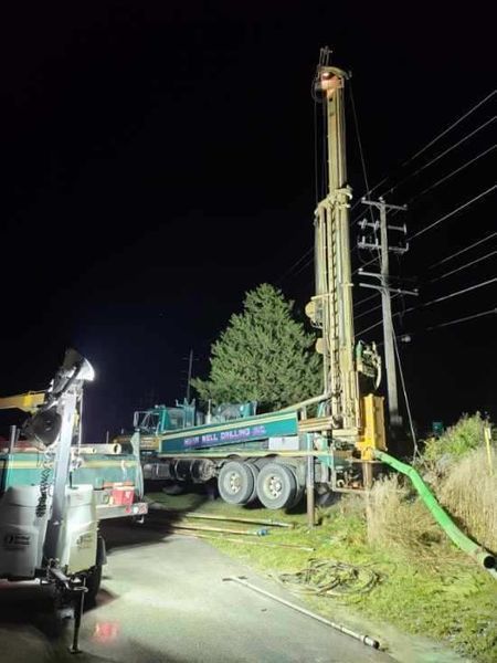 Drilling rig at night next to a power pole and greenery; green and white truck.