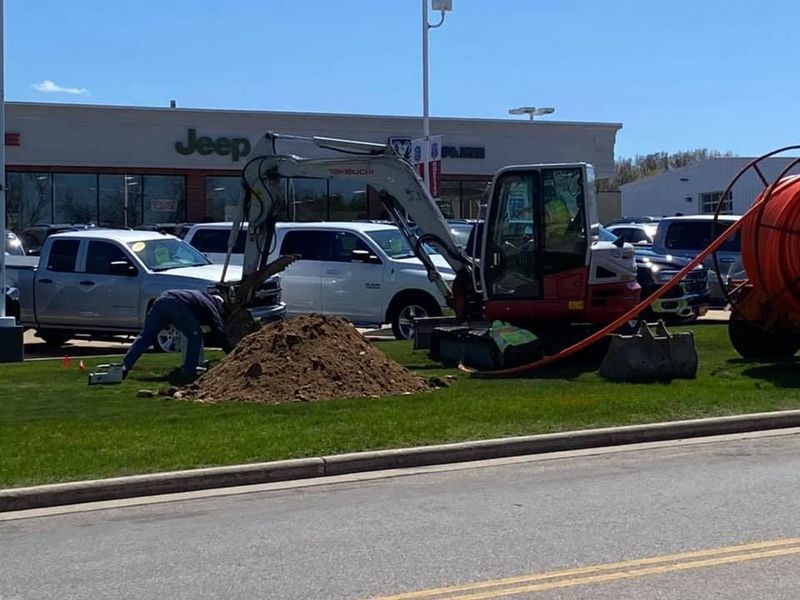 Construction worker and excavator digging on a lawn in front of a Jeep dealership, orange cable visible.