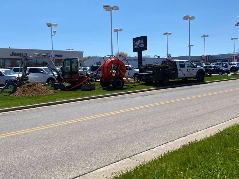 Construction site at a car dealership. An excavator and equipment trailer are on the grass beside a road. Sunny day.