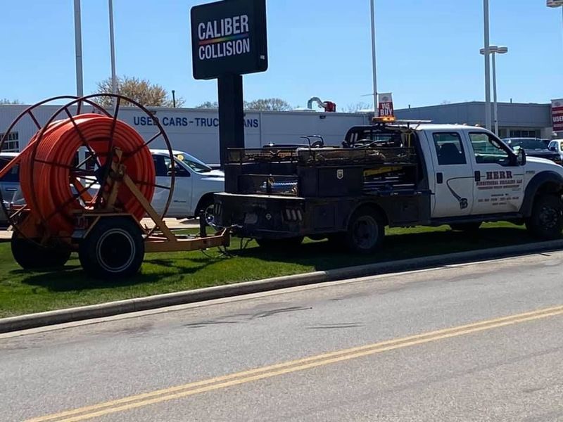 White truck with flatbed trailer hauling a large spool of orange conduit parked in front of a collision shop.