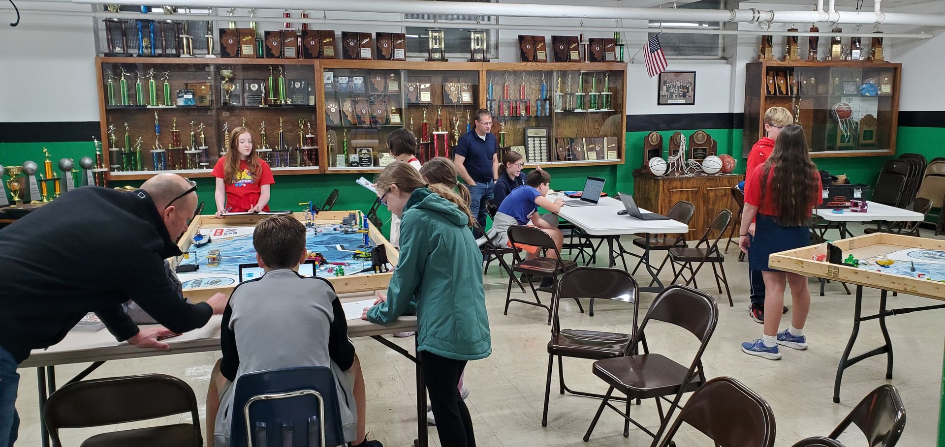 People at tables, a group activity in a room with trophies. Green and red accents.