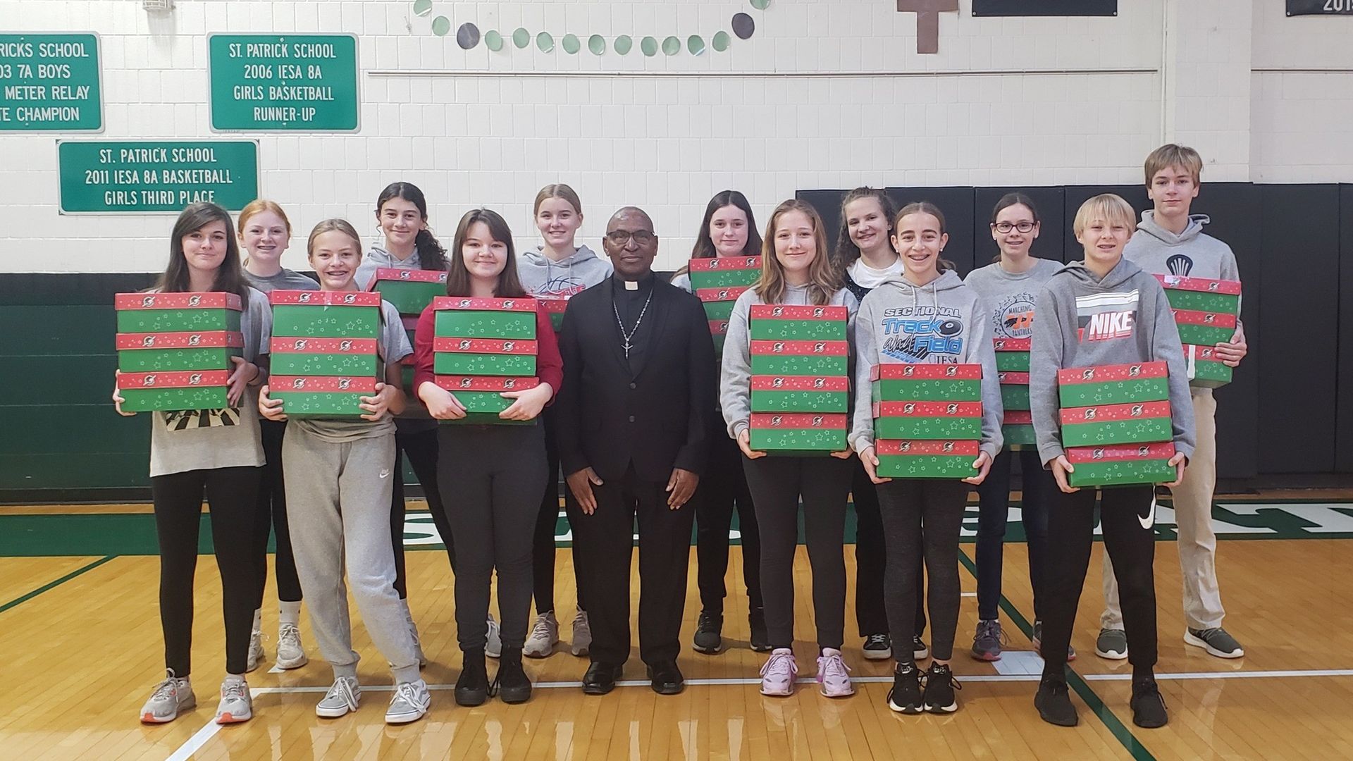 Students with gifts pose with a priest in a gym. They stand by a green wall with banners.
