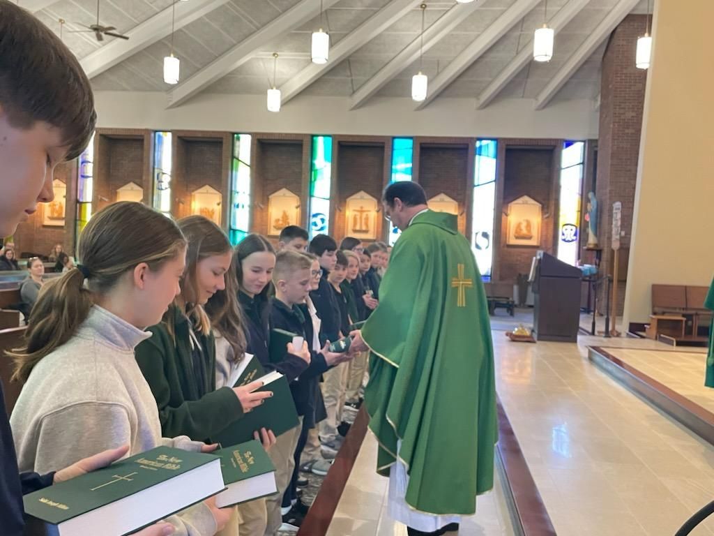 A priest in green vestments distributes books to students at a church altar.