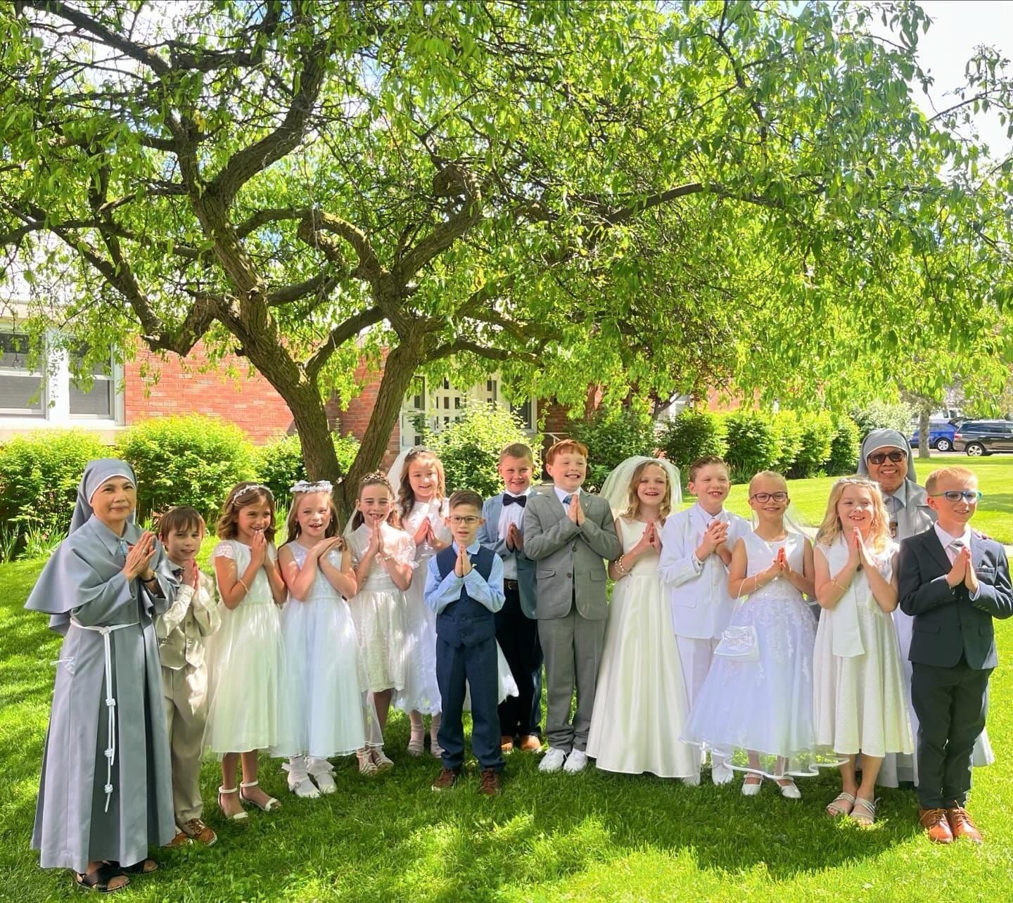 Children in white dresses and suits with clasped hands, with a nun, under a tree.