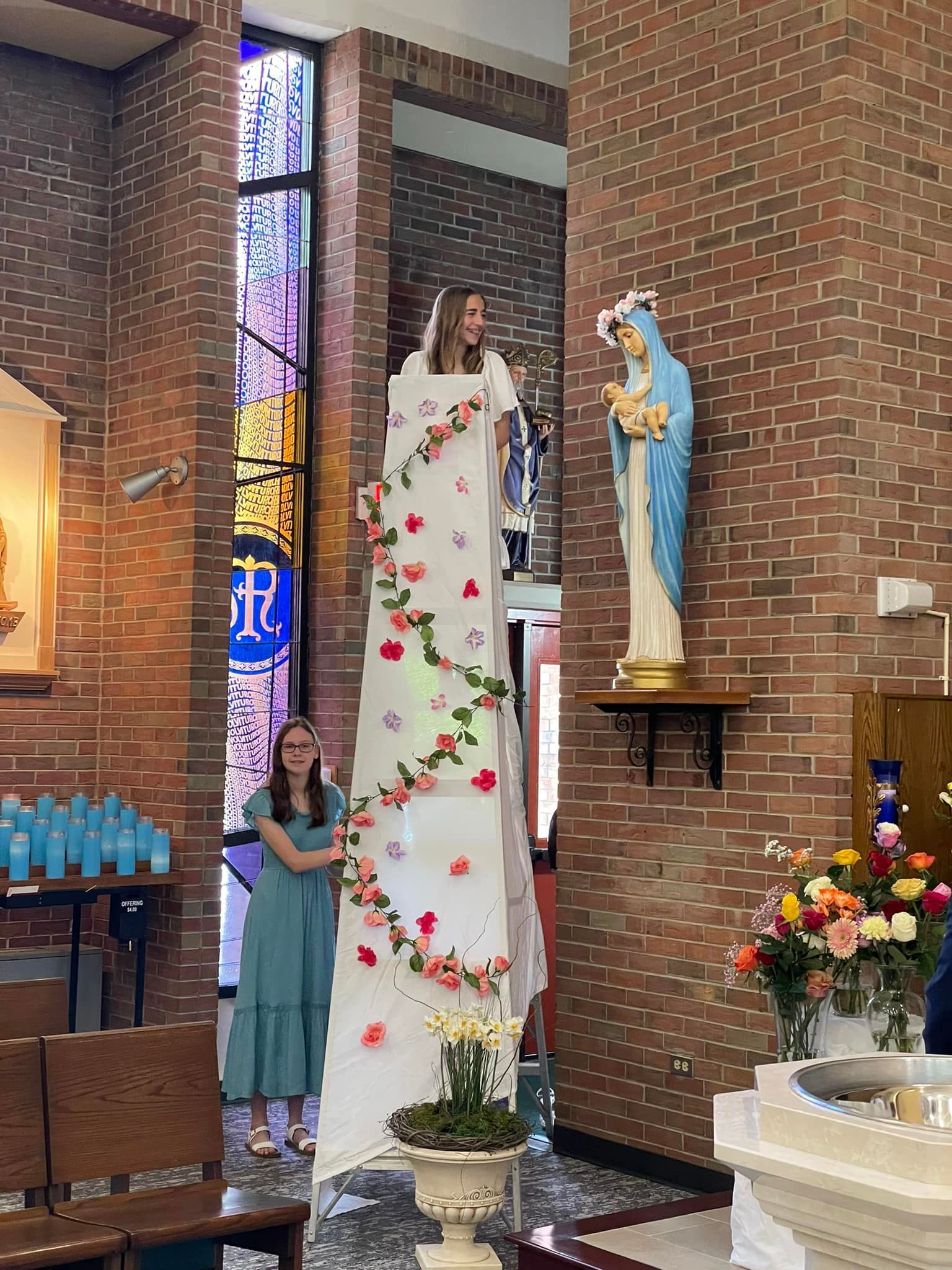 Two women beside a towering, flower-decorated paper dress inside a brick church; statue of Mary.