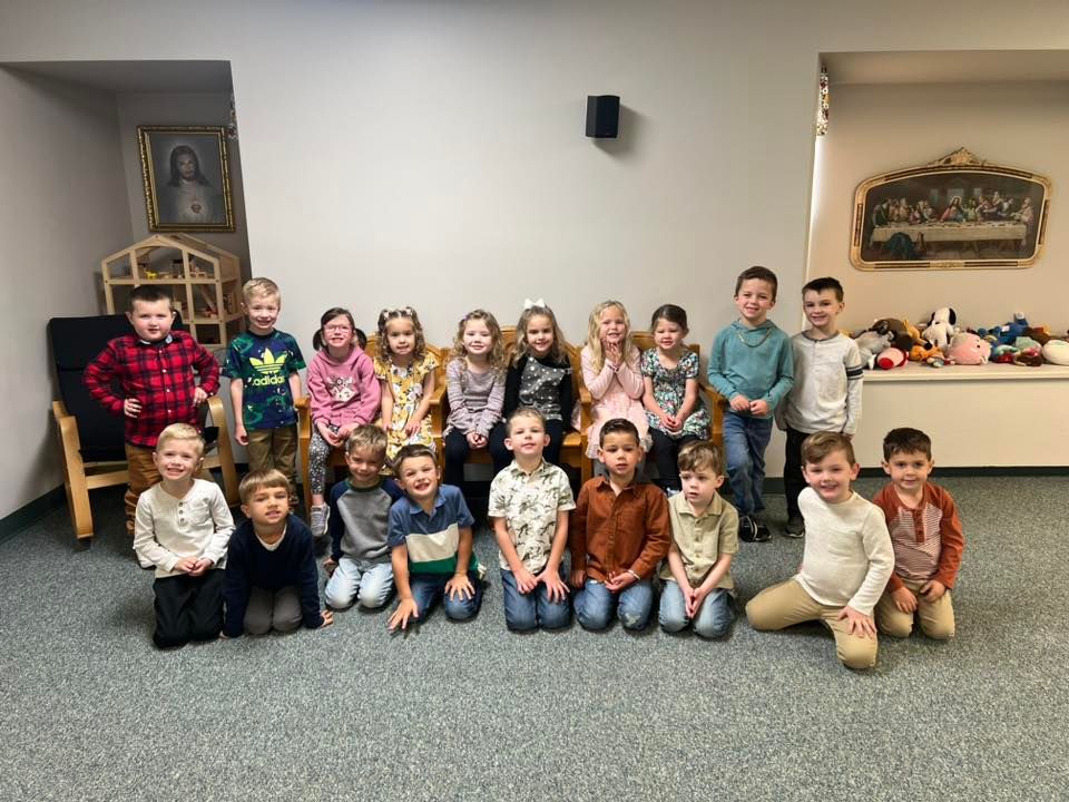 Group of young children, some kneeling, smiling, in a room with religious art.