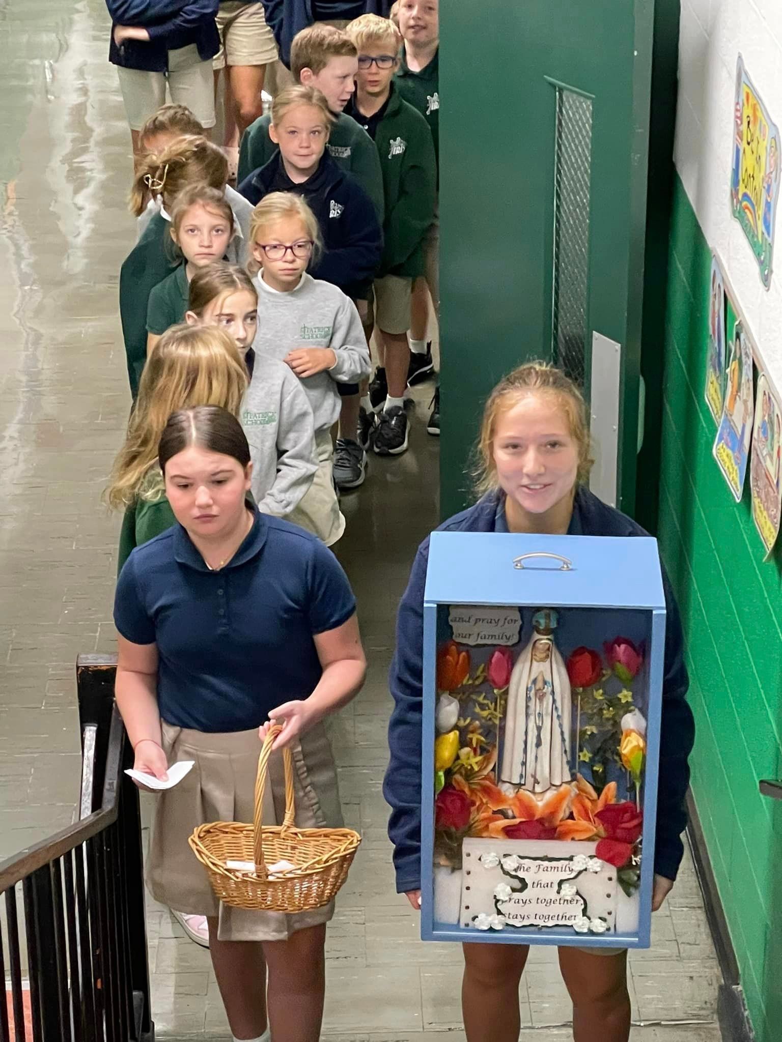Children in uniforms walking in line, one carrying a religious display box, others a basket, hallway.