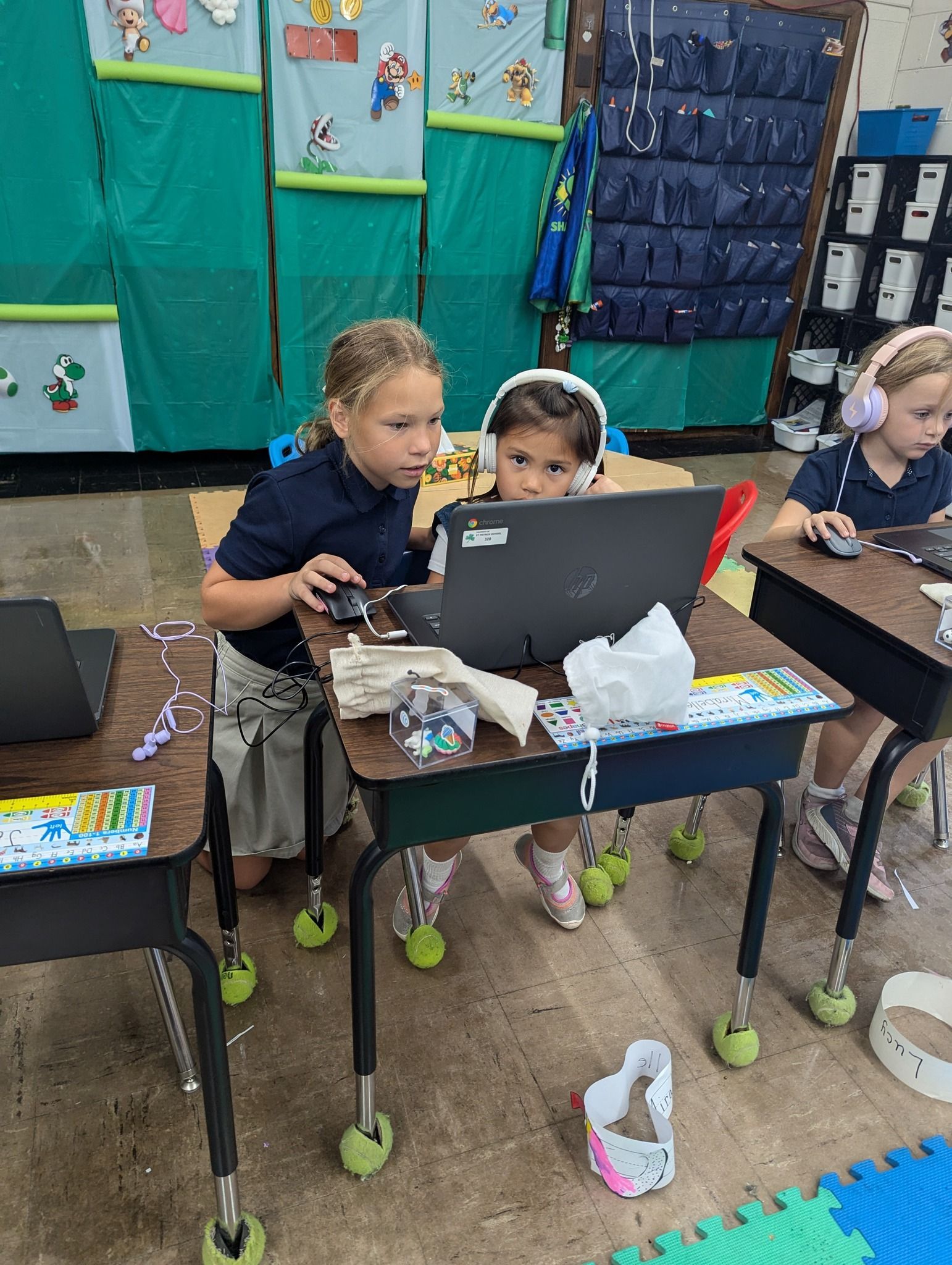 Three young students using laptops in a classroom. One student holds a mouse.
